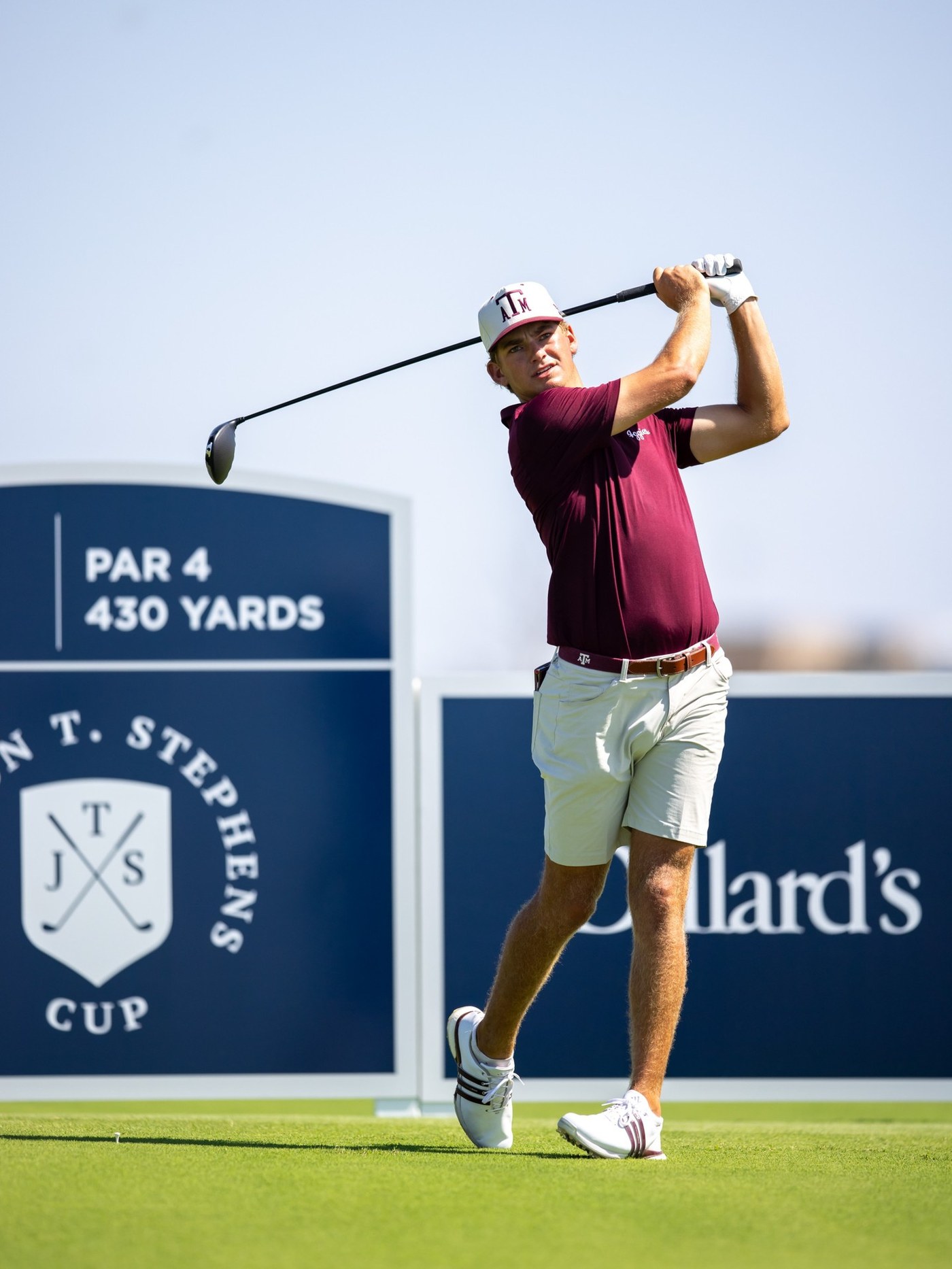 A Texas A&M golfer tees off at the Jackson T. Stephens Cup, with hole signage and the JTS Cup shield in the foreground